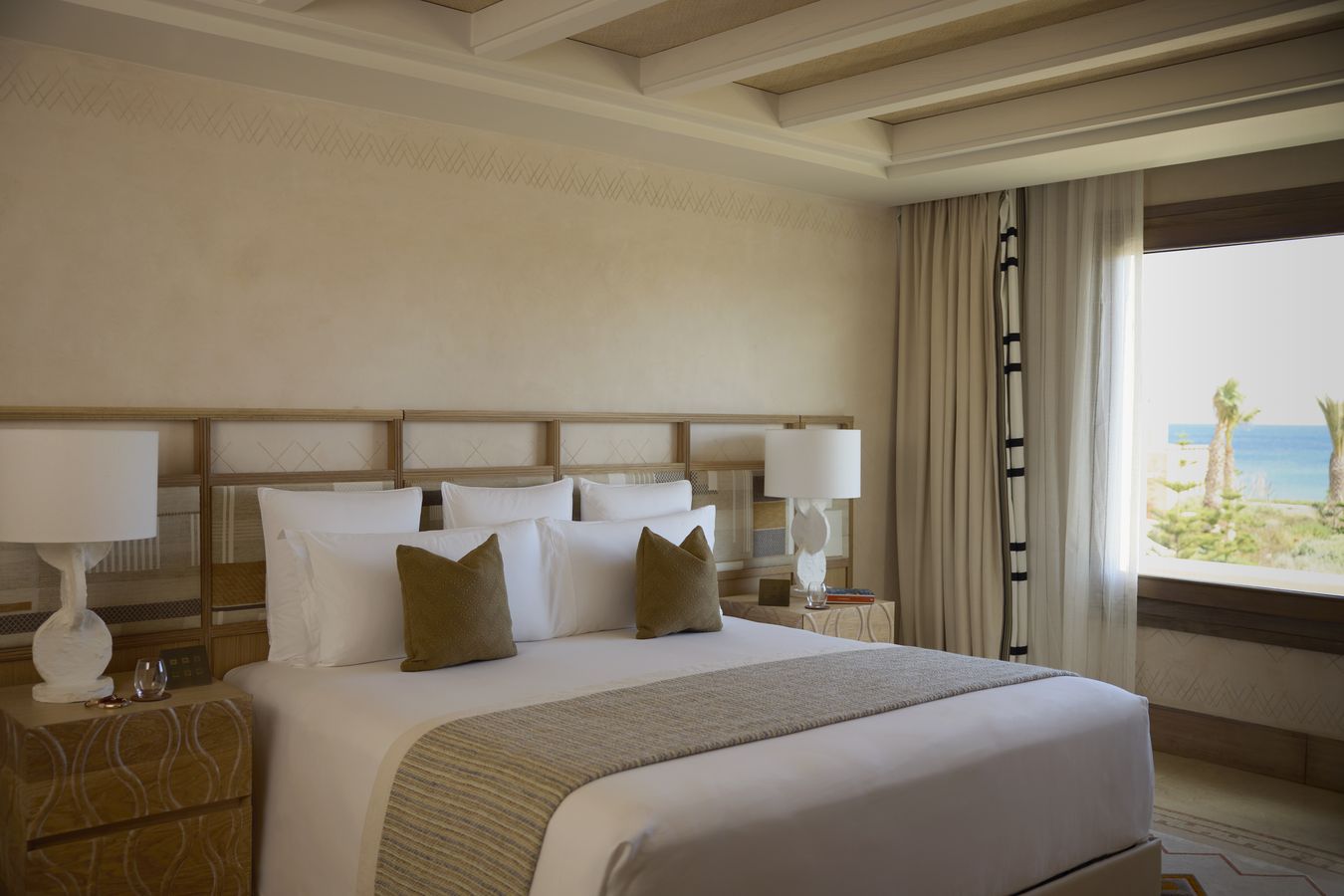 Lichte slaapkamer met groot bed en een raam met zicht op palmbomen en de zee in Royal Mansour Tamuda Bay